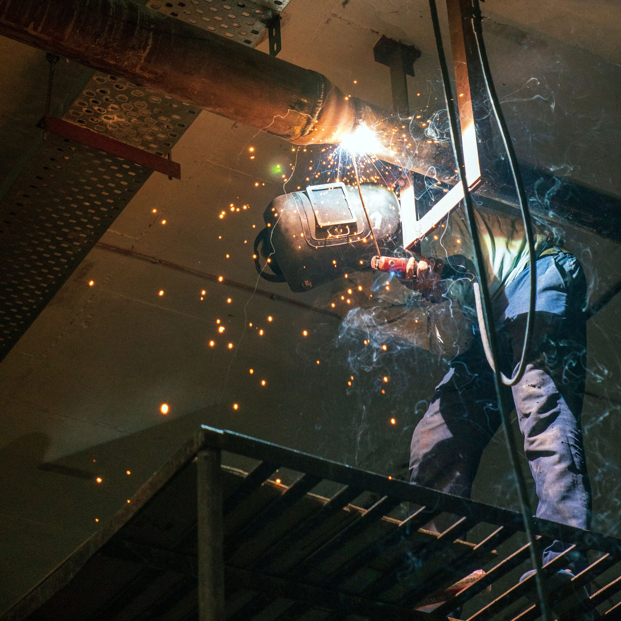 A welder working on metal pipes indoors, emitting bright sparks from the welding process.