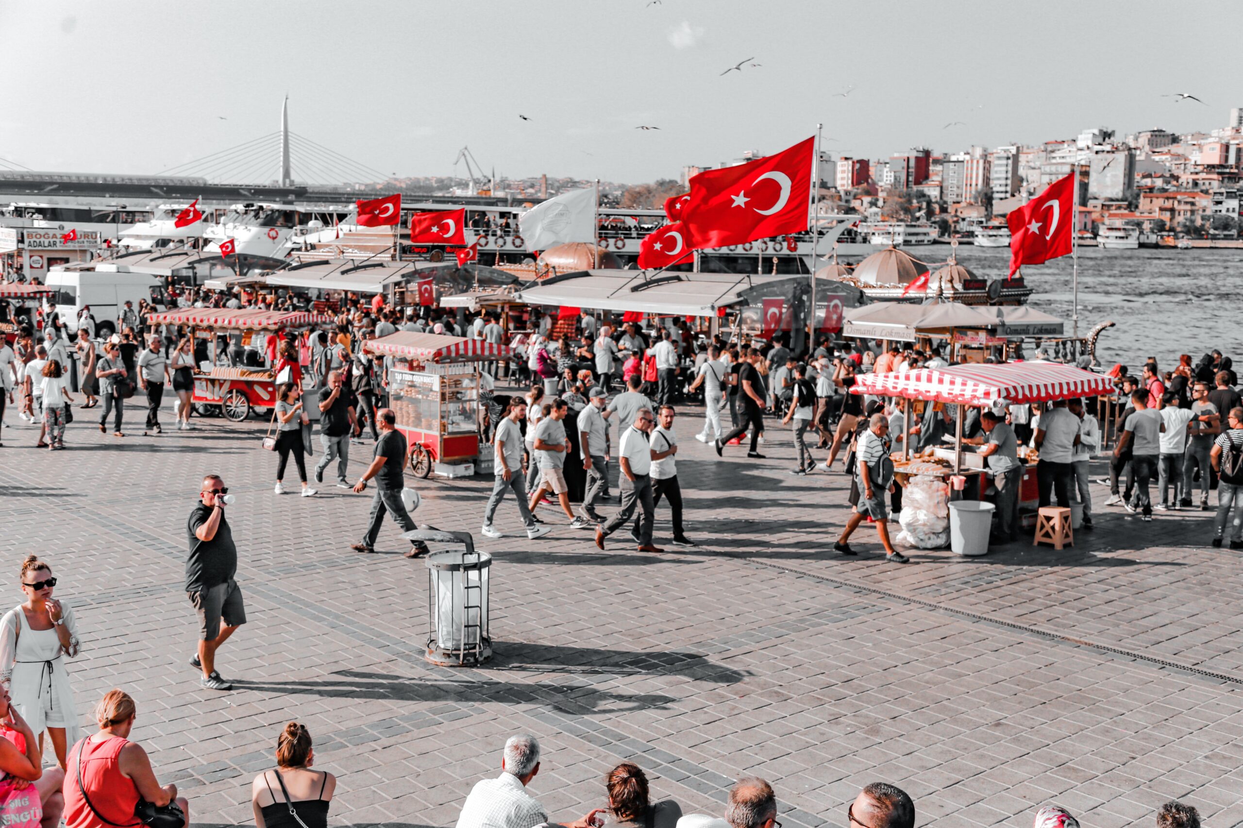 Busy street market in Istanbul with Turkish flags and bustling crowds by the riverside.